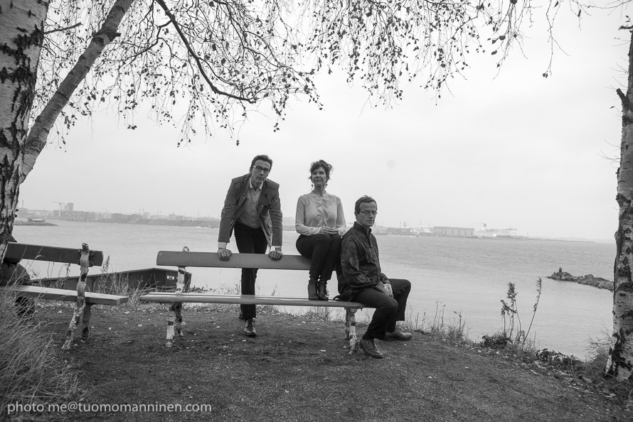 Kolme henkilöä istuu ja seisoo penkin äärellä puistossa merenrannalla.