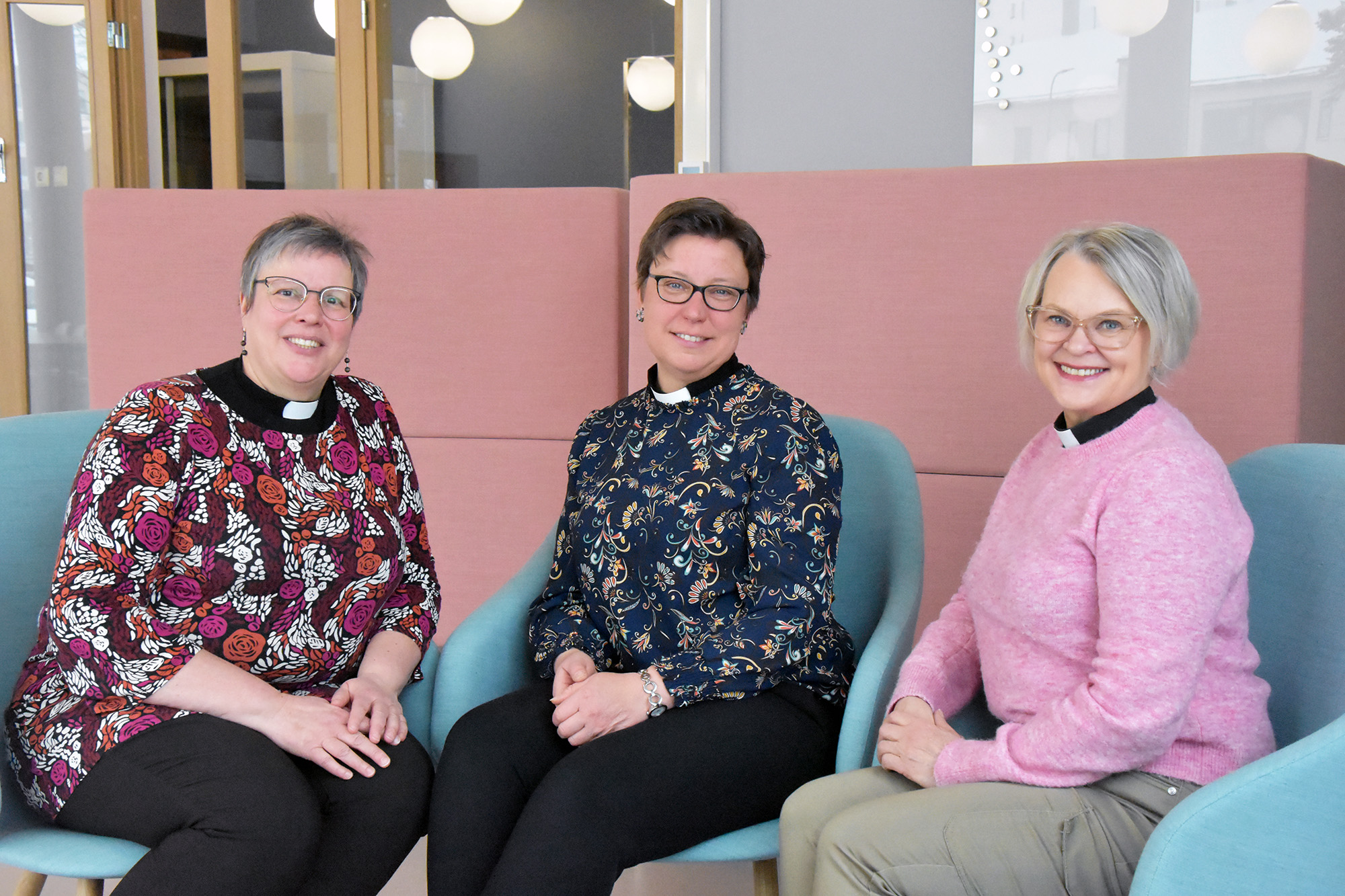 Kolme naista joilla on pappiskaulus, istuvat nojatuoleissa.