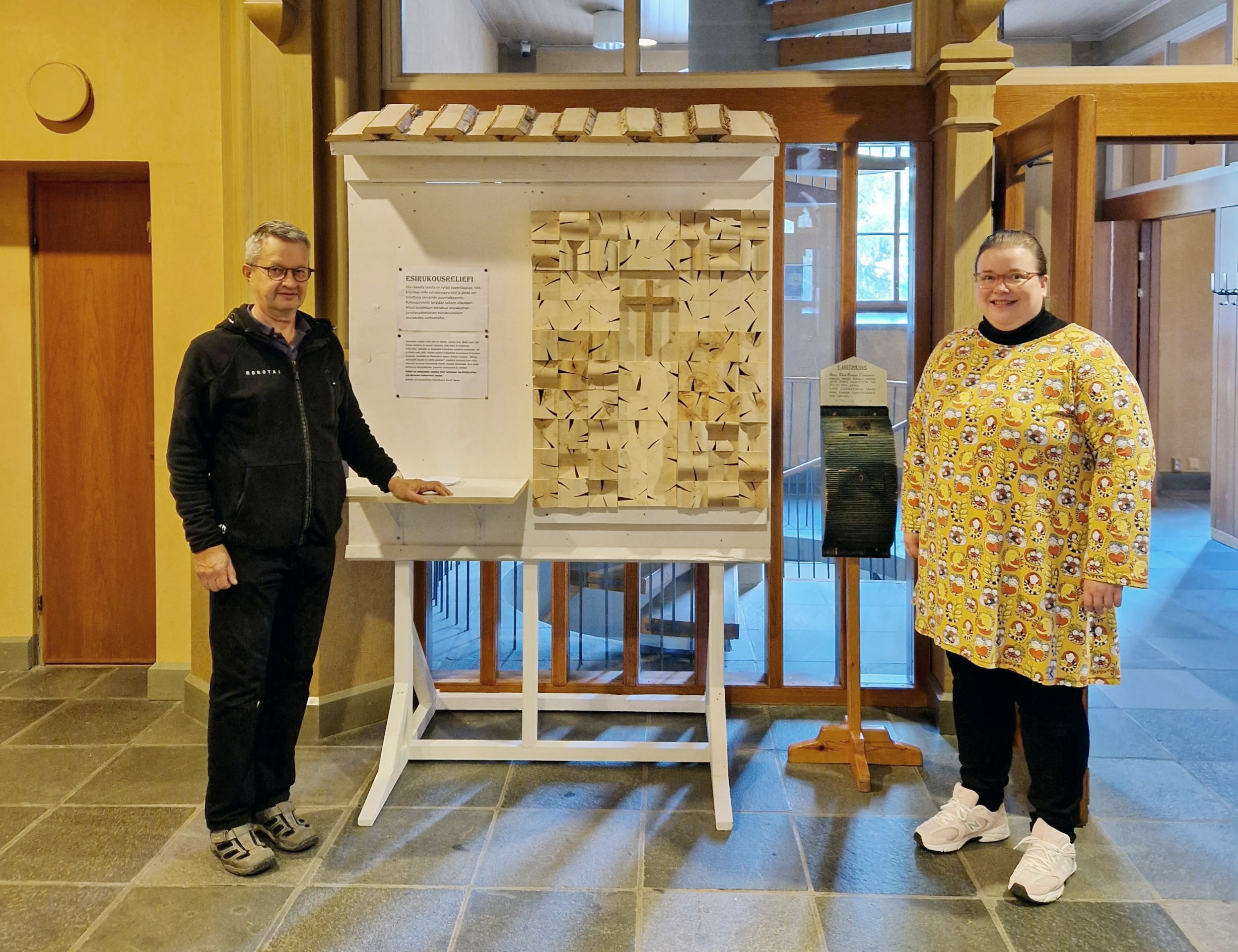 Mies ja nainen aulassa. heidän välissään on puinen reliefi ja lipas.