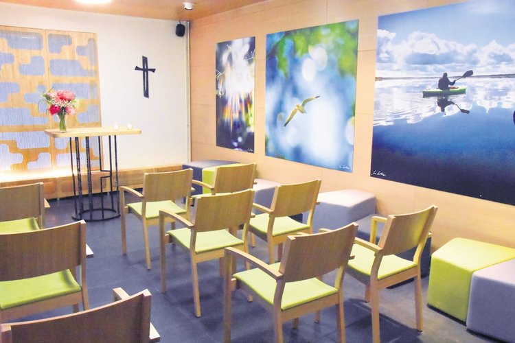 Hiljaisen huoneen seinillä on kauniita valokuvia ja risti. Huoneessa on vihreitä tuoleja rivissä.